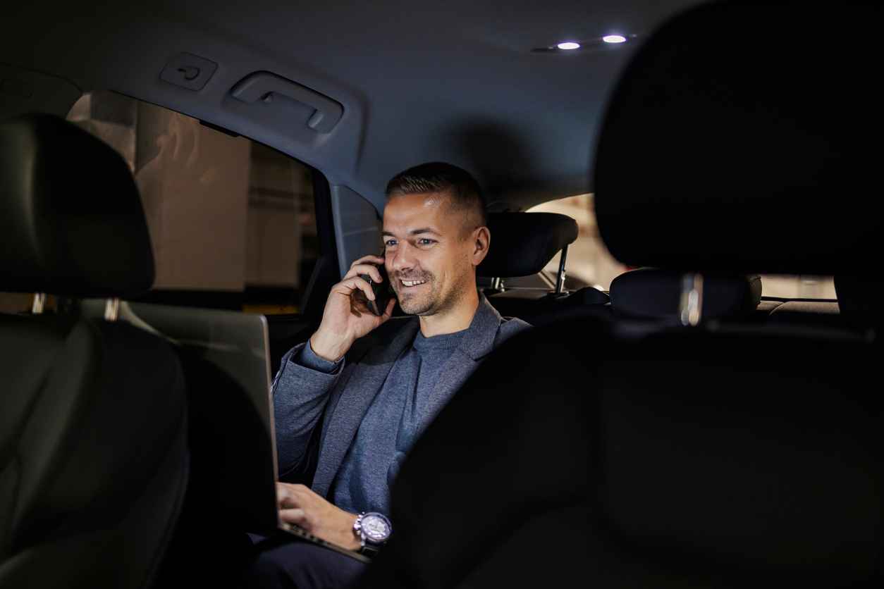Smiling busy executive on backseat in a car is having phone conversation and working on a laptop.